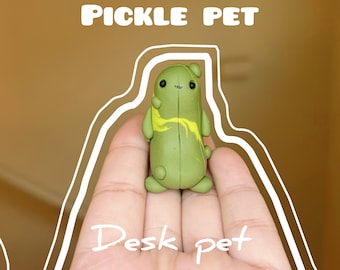 Puede incluir: Una figurita verde y amarilla con forma de pepinillo con una cara linda, etiquetada como "Pickle Pet" y "Desk Pet".