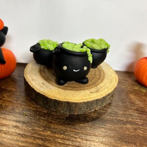 Peut inclure: Trois figurines de chaudrons noirs avec une potion verte bouillonnante, chacune avec un visage souriant. Les chaudrons sont pos&eacute;s sur une tranche de bois, avec une citrouille et une figurine de chat noir en arri&egrave;re-plan. D&eacute;cor sur le th&egrave;me d'Halloween.