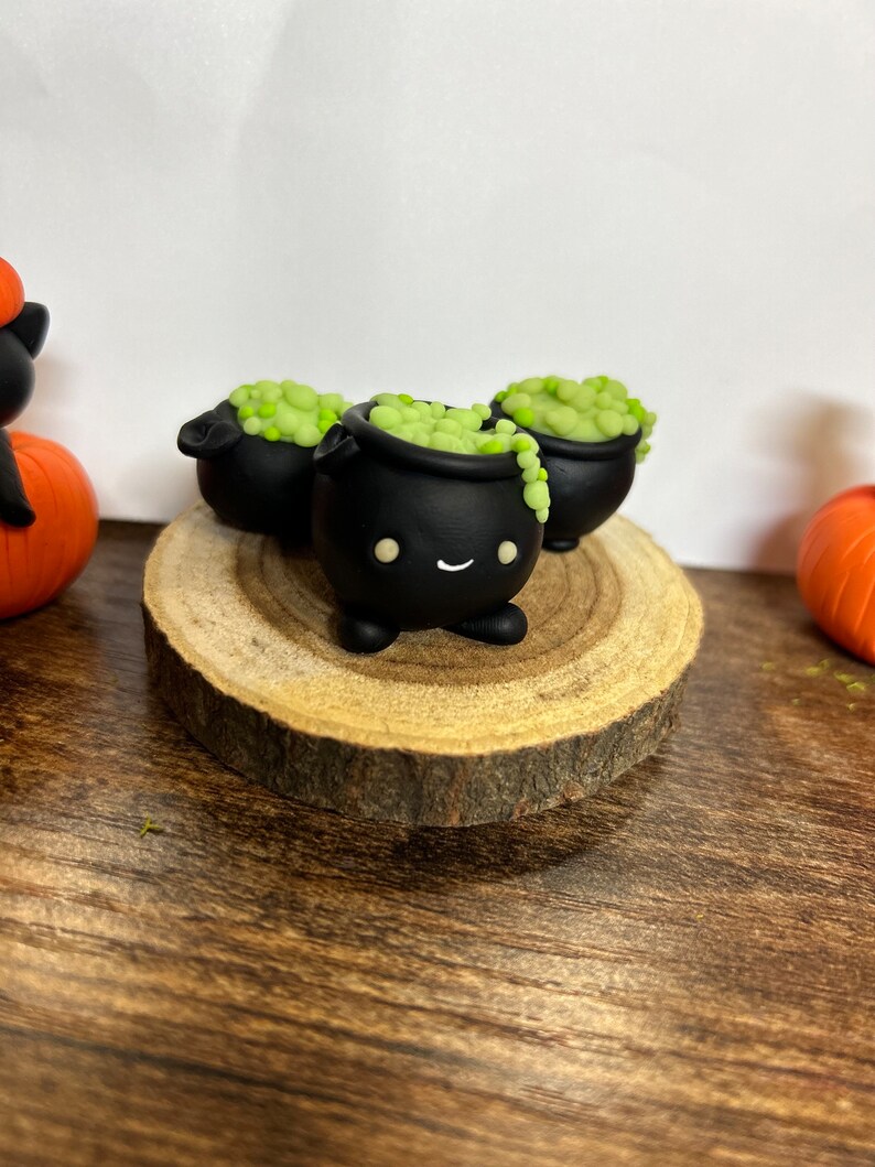 Peut inclure: Trois figurines de chaudrons noirs avec une potion verte bouillonnante, chacune avec un visage souriant. Les chaudrons sont pos&eacute;s sur une tranche de bois, avec une citrouille et une figurine de chat noir en arri&egrave;re-plan. D&eacute;cor sur le th&egrave;me d'Halloween.