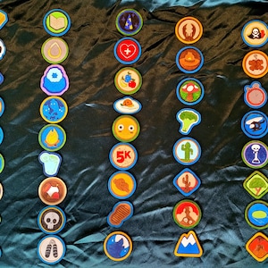 May include: A collection of colorful, circular patches with various designs, including a gold bar, a book, a wizard's hat, a heart, a skull, and a mountain. Each patch has a blue border and is arranged on a dark teal fabric.