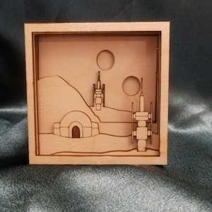 Peut inclure: Une boîte à ombre en bois avec un motif découpé au laser représentant un paysage désertique avec deux tours et un bâtiment en forme de dôme. La scène se déroule sur fond de deux lunes.