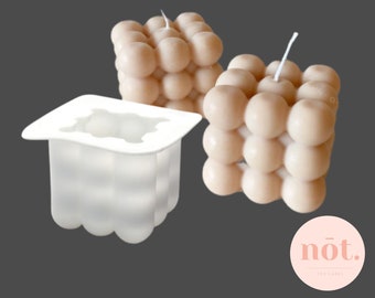 May include: Two beige 3D bubble candles and a white silicone mould for making bubble candles. The mould is square shaped with a rounded top and bottom. The candles are made of wax and have a white wick. The text "Silicone 3D Bubble Candle Molds" is at the top of the image. The text "nót. The Candy" is at the bottom of the image.