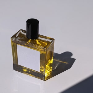 May include: A clear glass bottle with a black cap filled with a golden liquid. The bottle has a white label with a blank space for text.