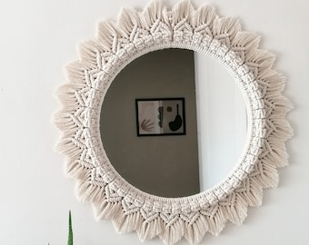 May include: A round mirror with a white macrame frame. The frame has a fringe border and is made with a knotting technique. The mirror is reflecting a room with a picture on the wall.