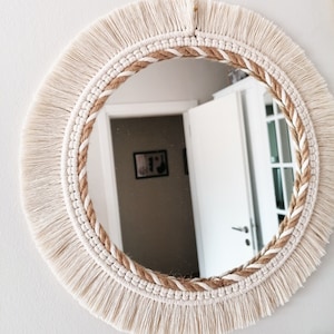 May include: A round mirror with a white fringe border and a natural rope design. The mirror is reflecting a room with white doors and a picture on the wall.