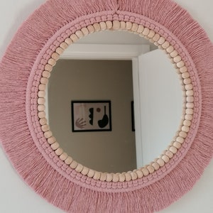 May include: A round mirror with a pink macrame frame and wooden beads. The mirror is hanging on a white wall.
