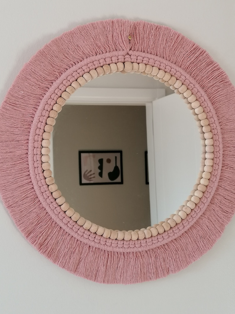May include: A round mirror with a pink macrame frame and wooden beads. The mirror is hanging on a white wall.