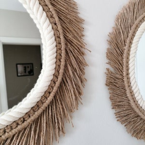 Op de afbeelding: Twee ronde spiegels met een bohemian design. De spiegels hebben een witte touwrand, een geweven bruine rand en een gefranjerde jute buitenrand. De spiegels zijn op een witte muur gemonteerd.