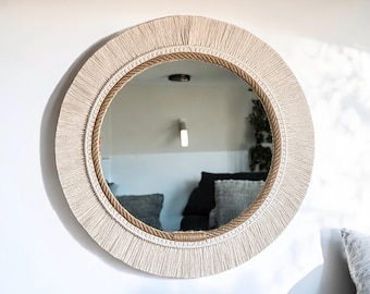 Może przedstawiać: Okrągłe lustro ścienne o naturalnym, boho designie. Lustro ma grubą, kremową frędzlę, sznurkowy detal i odblaskową szklaną powierzchnię. Neutralne odcienie i teksturowane elementy tworzą stylowy, dekoracyjny element wystroju wnętrz.