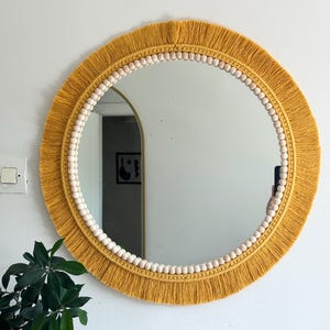 May include: A round wall mirror with a mustard yellow macrame fringe. The mirror's edge is adorned with small, light-colored wooden beads. The mirror is hanging on a white wall, with a glimpse of a plant in a pot.