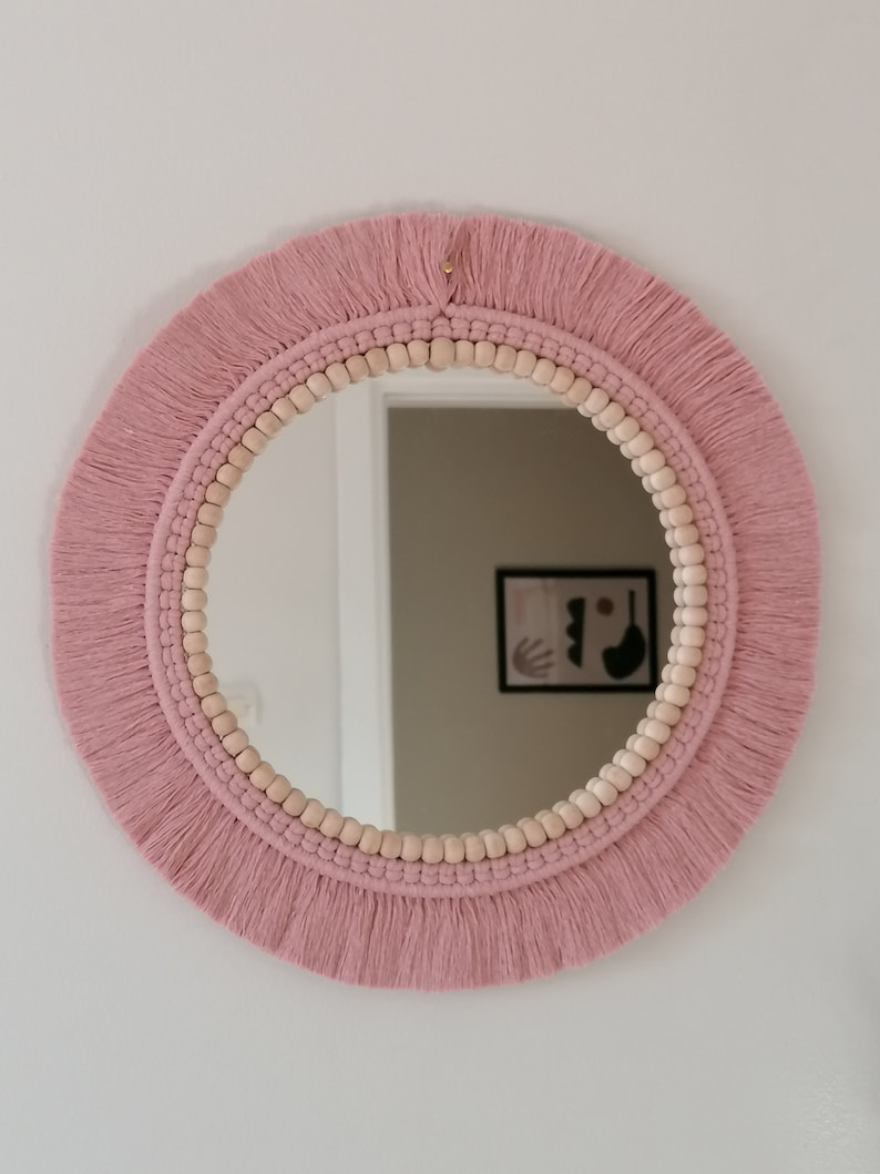 May include: A round mirror with a pink macrame frame and wooden beads. The mirror has a fringe border.