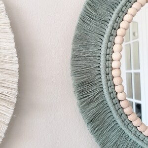 May include: A round mirror with a light green macrame frame and wooden beads. The mirror has a white background.