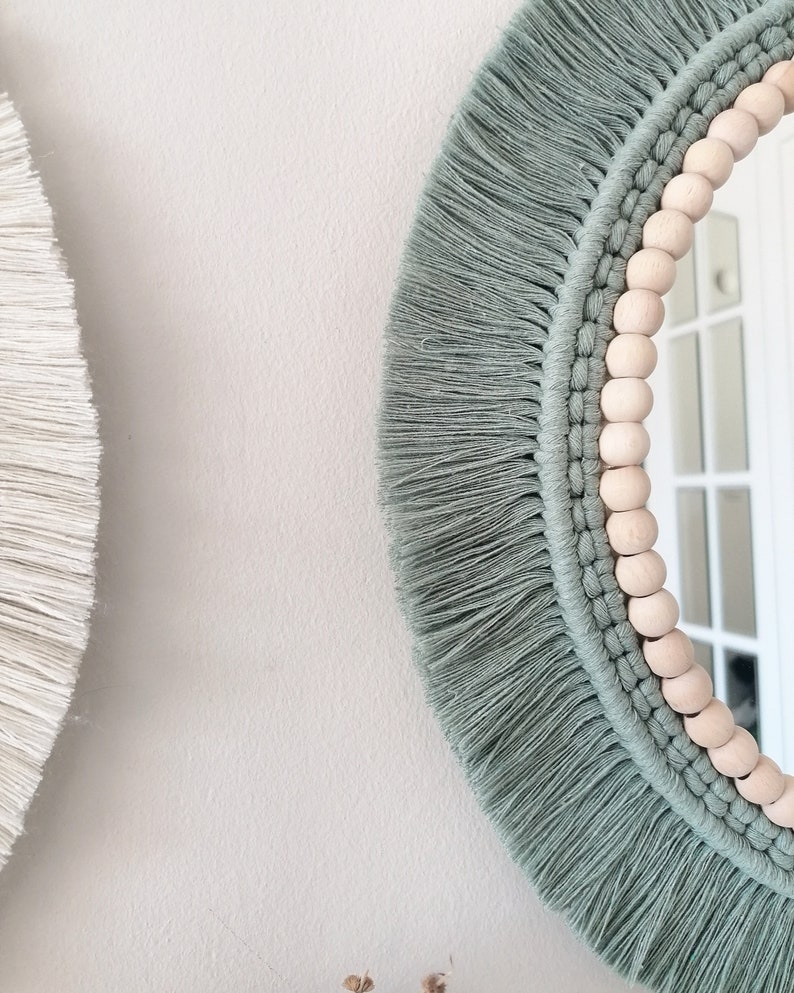 May include: A round mirror with a light green macrame frame and wooden beads. The mirror has a white background.