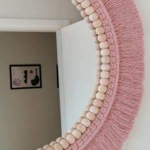 May include: A round mirror with a pink macrame frame and wooden beads. The mirror is surrounded by a fringe of pink yarn.