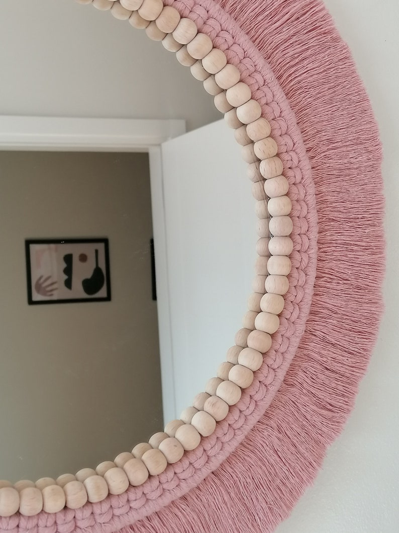 May include: A round mirror with a pink macrame frame and wooden beads. The mirror is surrounded by a fringe of pink yarn.
