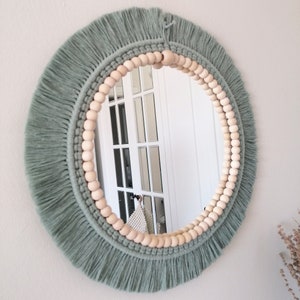 May include: A round mirror with a sage green macrame frame and a wooden bead border. The mirror is reflecting a white door with glass panels.