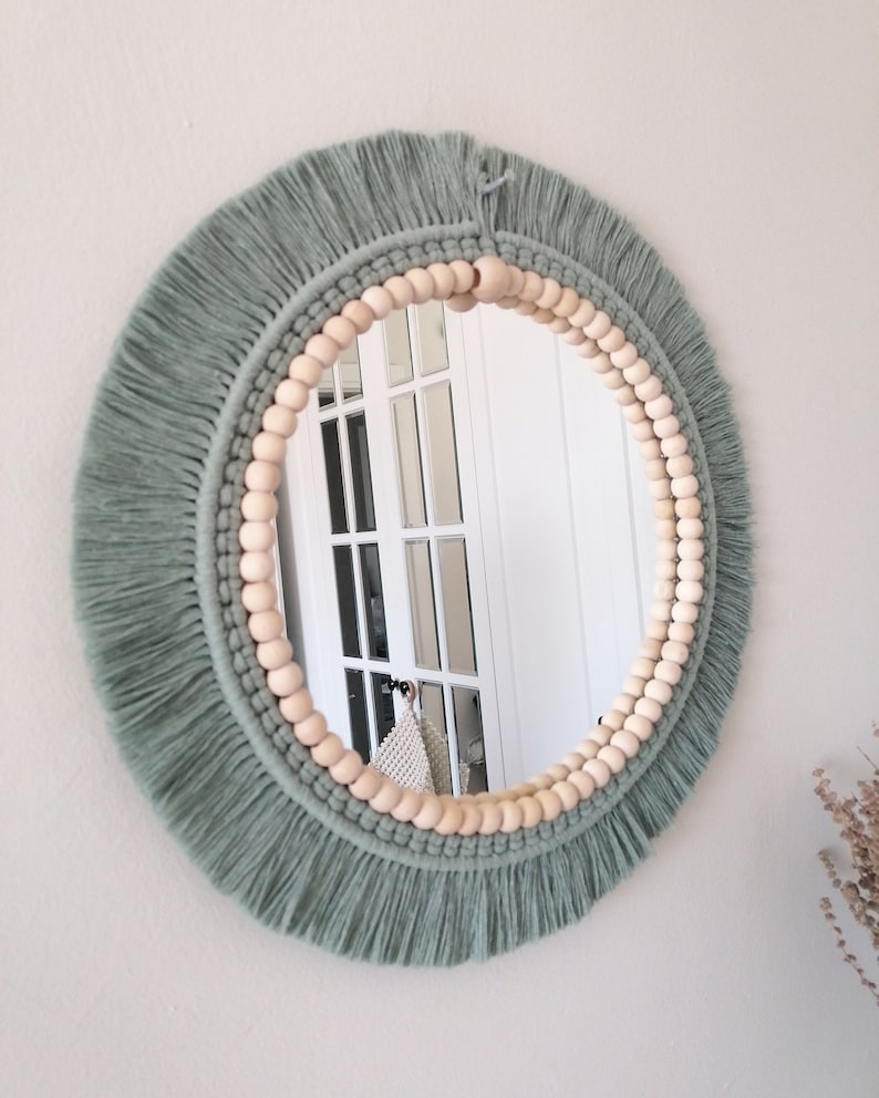 May include: A round mirror with a sage green macrame frame and a wooden bead border. The mirror is reflecting a white door with glass panels.