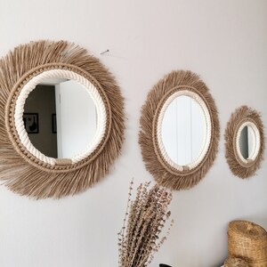 Op de afbeelding: Drie ronde spiegels met een bohemian design. Elke spiegel heeft een franje van natuurlijke vezels en een witte touwrand. De spiegels zijn verschillend van grootte en zijn bevestigd aan een lichtgrijze muur. Een bos gedroogde bloemen en decoratieve manden staan op de voorgrond.