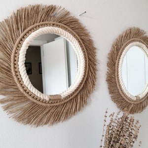Op de afbeelding: Twee ronde spiegels met een natuurlijk, boheems ontwerp. De spiegels hebben een centraal reflecterend oppervlak, omgeven door een dik, cr&egrave;mekleurig touw en een gefranjerde, geweven rand in lichtbruin. Ze zijn aan een witte muur bevestigd.