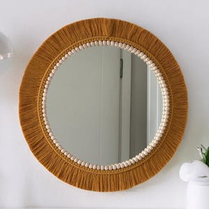 May include: A circular macrame mirror in a mustard hue, featuring a fringed edge and a beaded inner ring. This decorative wall hanging offers a bohemian aesthetic for home interiors. The mirror's diameter is approximately 50 cm.