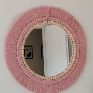 May include: A round mirror with a pink macrame frame and wooden beads. The mirror has a simple design and would look great in a modern or bohemian home.