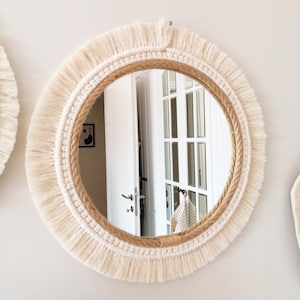 May include: A round mirror with a white macrame frame and a natural rope border. The mirror is reflecting a white door with glass panels.