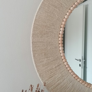 May include: A round mirror with a natural fiber wrapped frame. The frame is beige and has a radial design. Small, light-colored wooden beads line the outer edge of the frame. The mirror is hanging on a white wall.