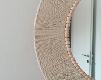 May include: A round mirror with a natural fiber wrapped frame. The frame is beige and has a radial design. Small, light-colored wooden beads line the outer edge of the frame. The mirror is hanging on a white wall.