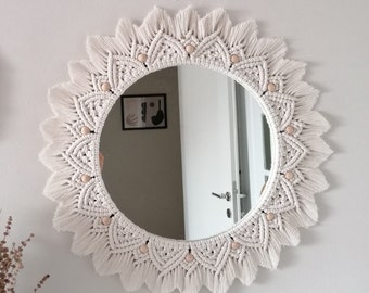 May include: A white macrame mirror with a circular shape and a fringe border. The mirror has a wooden bead design around the edge.