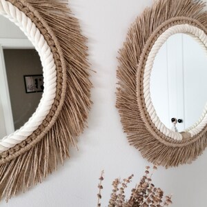 Op de afbeelding: Twee ronde spiegels met een natuurlijk, boheems ontwerp. Elke spiegel heeft een witte touwrand, een geweven bruin frame en een franje van bruine vezels. De spiegels zijn bevestigd aan een witte muur, met gedroogde bloemen op de voorgrond.
