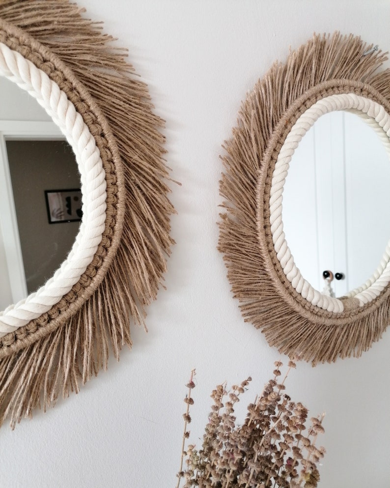 Op de afbeelding: Twee ronde spiegels met een natuurlijk, boheems ontwerp. Elke spiegel heeft een witte touwrand, een geweven bruin frame en een franje van bruine vezels. De spiegels zijn bevestigd aan een witte muur, met gedroogde bloemen op de voorgrond.