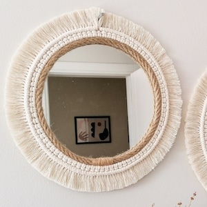 May include: A round mirror with a white macrame frame and a natural jute rope border. The mirror is hanging on a white wall.