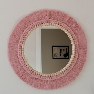 May include: A round pink mirror with a fringe border and a wooden bead trim. The mirror is hanging on a white wall.