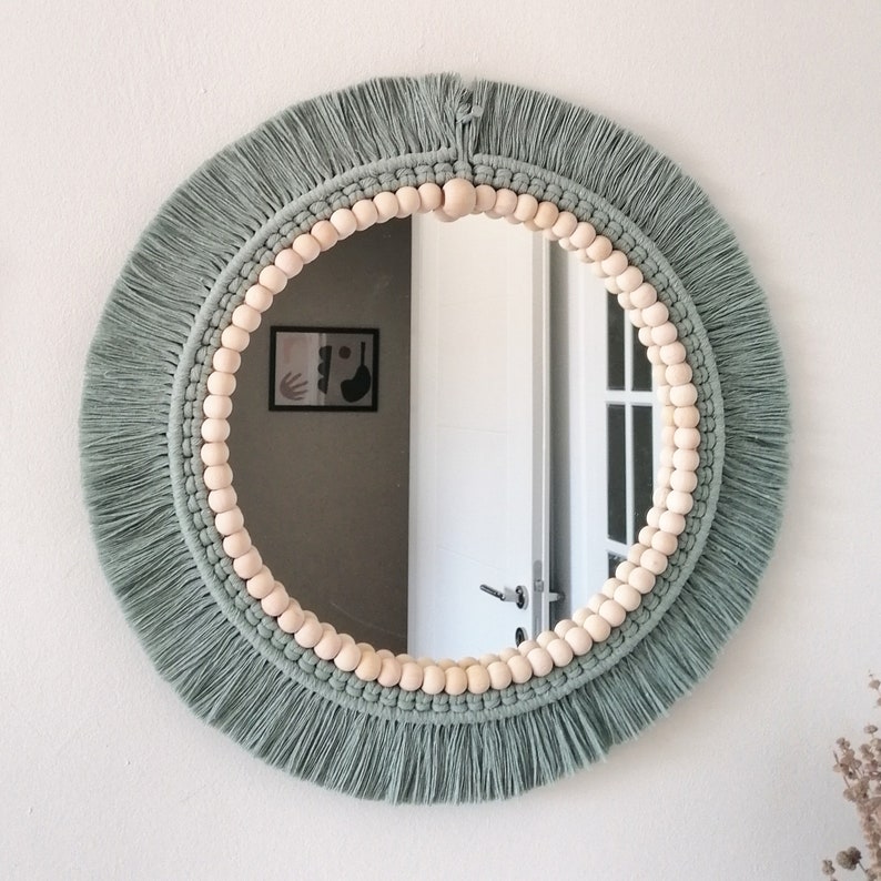 May include: A round mirror with a macrame frame and a border of wooden beads. The frame is a sage green color and the beads are a natural wood color.