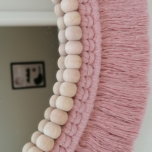 May include: A pink macrame mirror with a beaded border. The mirror is round and has a fringe around the edge.