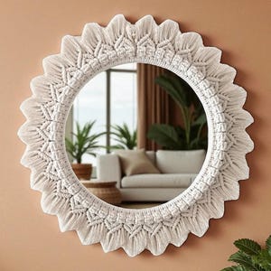 May include: Round white macrame mirror with a scalloped edge. The mirror's frame features intricate woven patterns and a fringe detail. The mirror is mounted on a peach-colored wall, reflecting a living room scene.