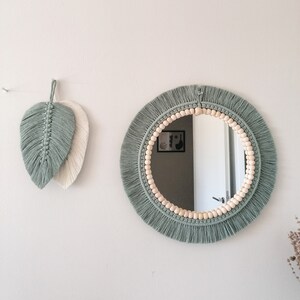 May include: A round mirror with a macrame frame and wooden beads. The frame is a sage green color and the beads are a natural wood color. The mirror is hanging on a white wall.