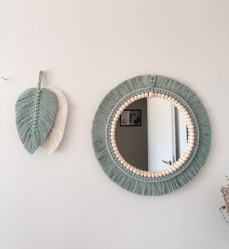 May include: A round mirror with a macrame frame and wooden beads. The frame is a sage green color and the beads are a natural wood color. The mirror is hanging on a white wall.