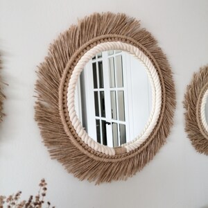 Op de afbeelding: Ronde wandspiegel met een natuurlijke jute franje en een cr&egrave;mekleurige touwrand. De spiegel is bevestigd aan een witte muur. Het ontwerp is bohemien en rustiek. De diameter van de spiegel is ongeveer 40 cm.