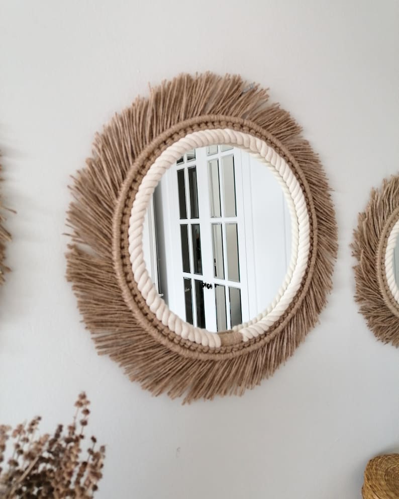Op de afbeelding: Ronde wandspiegel met een natuurlijke jute franje en een cr&egrave;mekleurige touwrand. De spiegel is bevestigd aan een witte muur. Het ontwerp is bohemien en rustiek. De diameter van de spiegel is ongeveer 40 cm.