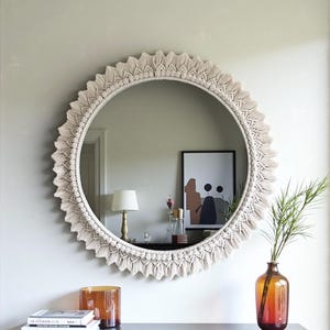 May include: A round mirror with a cream-colored macrame fringe. The mirror hangs on a light gray wall above a dark wood console table. A lamp, books, and a vase with greenery are on the table.