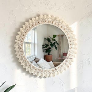 May include: Round mirror with a cream-colored macrame sunburst design. The mirror's frame is white, and the macrame features small, light brown beads. The mirror is hanging on a textured white wall, reflecting a room with a window and plants.