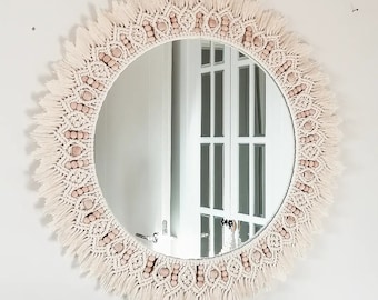Op de afbeelding: Een ronde spiegel met een macramé-frame en houten kralen. Het frame is crèmekleurig en heeft een franje rond de rand. De spiegel reflecteert een witte deur met glazen panelen.