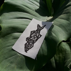 Könnte beinhalten: Ein kleines, rechteckiges Stoffpatch mit einem schwarzen Fischdesign. Das Patch befindet sich auf einem großen, grünen Blatt mit ausgeprägten Adern. Die Fischillustration ist detailliert und stilisiert.