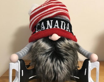 Może przedstawiać: Ozdobny gnom w czerwono-białej czapce w paski z białym pomponem, czarnej czapce z napisem "CANADA" w białych literach oraz długiej szarej i czarnej brodzie. Gnom siedzi na czarno-białych łyżwach.