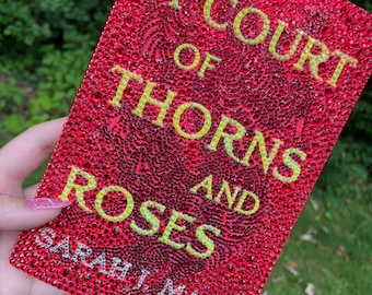 May include: A red book cover with a title that reads "A Court of Thorns and Roses" in yellow lettering. The author's name, "Sarah J. Maas", is printed in white lettering at the bottom of the cover.