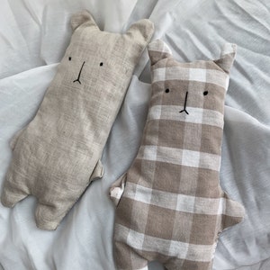 Könnte beinhalten: Zwei Plüschkissen in Bärenform. Eines ist beige mit einem einfachen gestickten Gesicht, das andere ist beige und weiß kariert mit einem ähnlichen gestickten Gesicht.