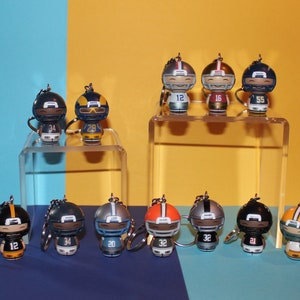 May include: A collection of miniature football player keychains. Each keychain features a stylized figure wearing a helmet and jersey with a unique number. The keychains are displayed on a clear acrylic stand against a blue and yellow backdrop.