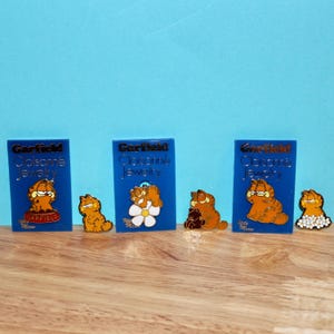 May include: A collection of Garfield cloisonné jewelry pins. The pins feature the orange cat in various poses, some with flowers or teddy bears. The pins are displayed on blue cards with the Garfield logo and "Colsonne Jewelry" text.