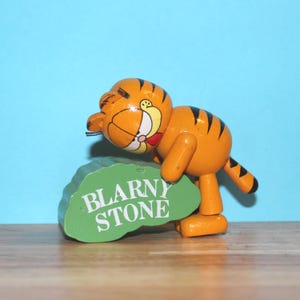 May include: Wooden orange tiger figurine with black stripes, holding a green sign that reads "BLARNY STONE". The tiger has a yellow face with a red bow tie and is posed on a wooden surface against a blue background.
